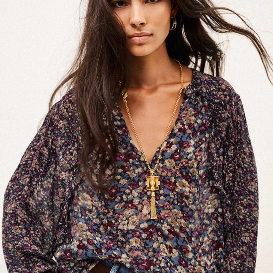 A woman modeling a long-sleeve V-neck blouse with a dense multicolor floral print in purple, blue, beige, and green, styled with blue jeans and a gold pendant necklace against a plain white background.
