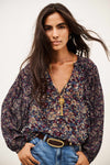 A woman modeling a long-sleeve V-neck blouse with a dense multicolor floral print in purple, blue, beige, and green, styled with blue jeans and a gold pendant necklace against a plain white background.