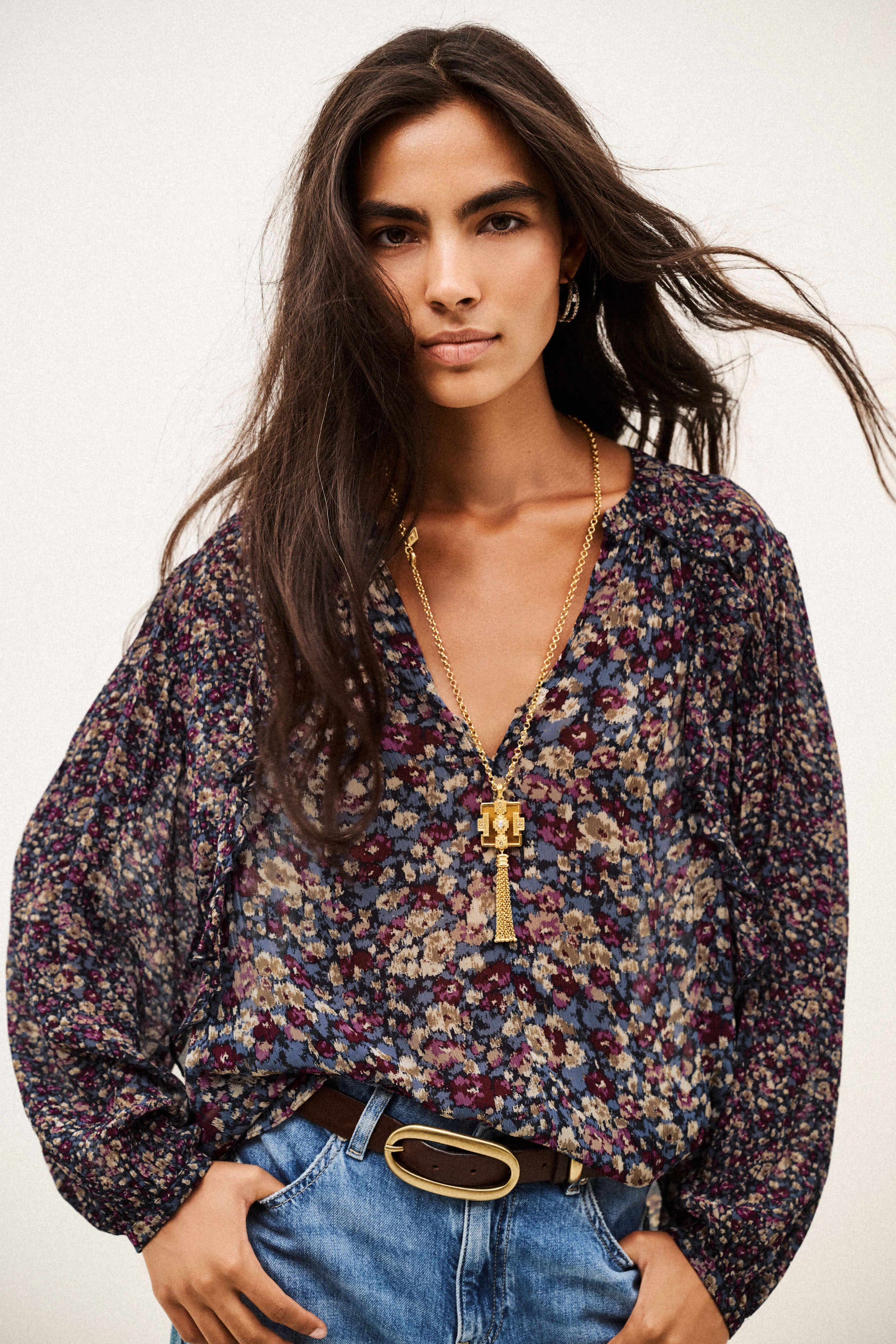 A woman modeling a long-sleeve V-neck blouse with a dense multicolor floral print in purple, blue, beige, and green, styled with blue jeans and a gold pendant necklace against a plain white background.