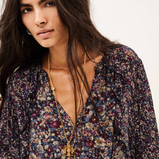 A woman wearing a long-sleeve bohemian floral print blouse with a deep V-neckline, layered with gold necklaces, against a plain off-white background.