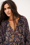 A woman wearing a long-sleeve bohemian floral print blouse with a deep V-neckline, layered with gold necklaces, against a plain off-white background.