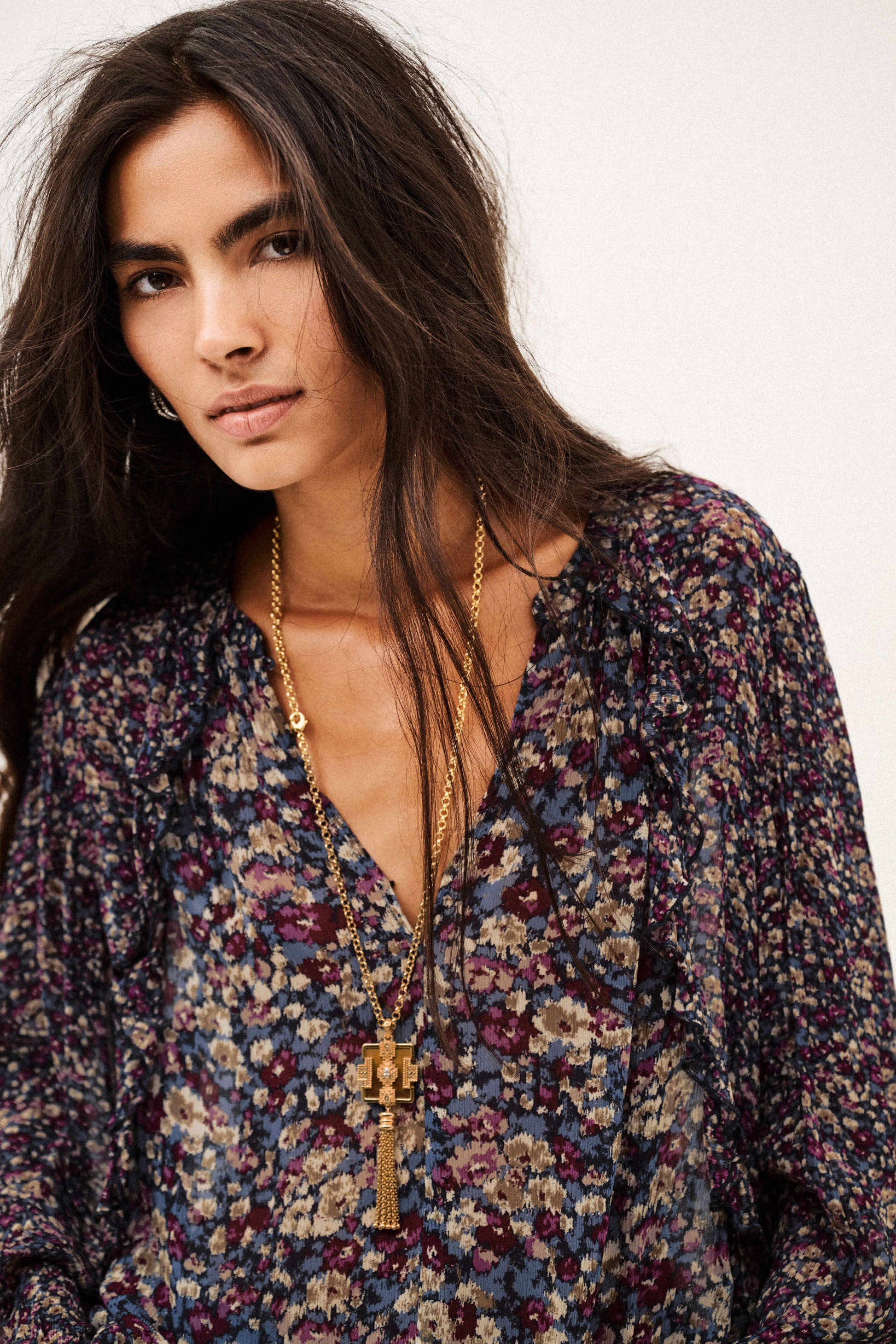 A woman wearing a long-sleeve bohemian floral print blouse with a deep V-neckline, layered with gold necklaces, against a plain off-white background.