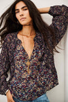 A woman wearing a long-sleeve bohemian floral print blouse with a deep V-neck, paired with a gold key pendant necklace and blue jeans, posing against a light gray wall.