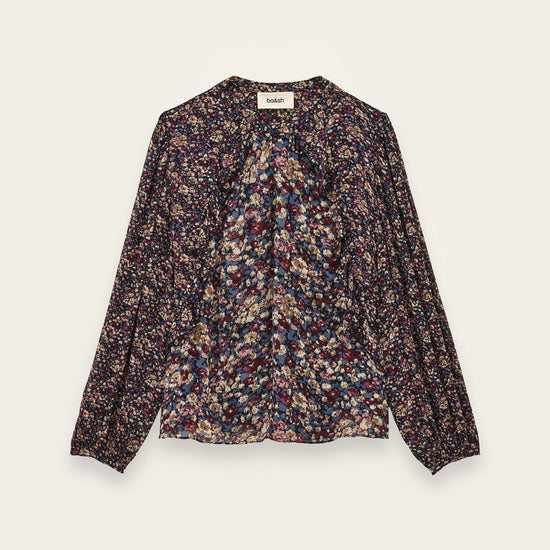 A long-sleeve blouse with a dense multicolor floral and abstract print on a dark base, featuring a gathered front and a round neckline, shown flat against a light cream background.