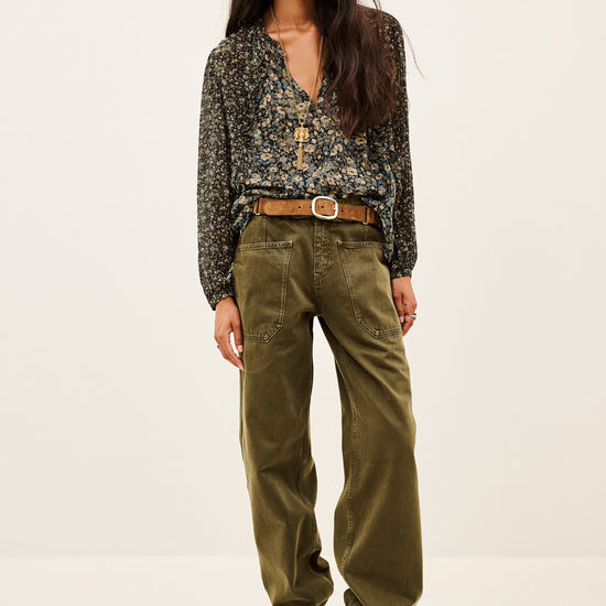 A woman modeling a dark floral long-sleeve blouse tucked into olive green wide-leg pants with a brown belt and brown ankle boots, photographed against a plain light studio background.
