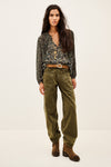 A woman modeling a dark floral long-sleeve blouse tucked into olive green wide-leg pants with a brown belt and brown ankle boots, photographed against a plain light studio background.
