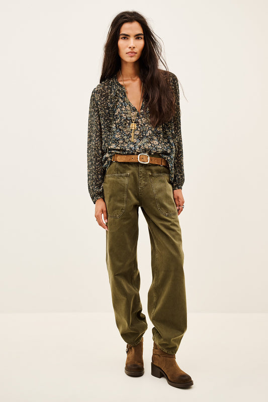 A woman modeling a dark floral long-sleeve blouse tucked into olive green wide-leg pants with a brown belt and brown ankle boots, photographed against a plain light studio background.