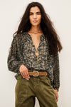 A woman modeling a long-sleeve floral blouse with a deep V-neck and a gold cross pendant, paired with olive green cargo pants and a brown belt, against a plain off-white background.