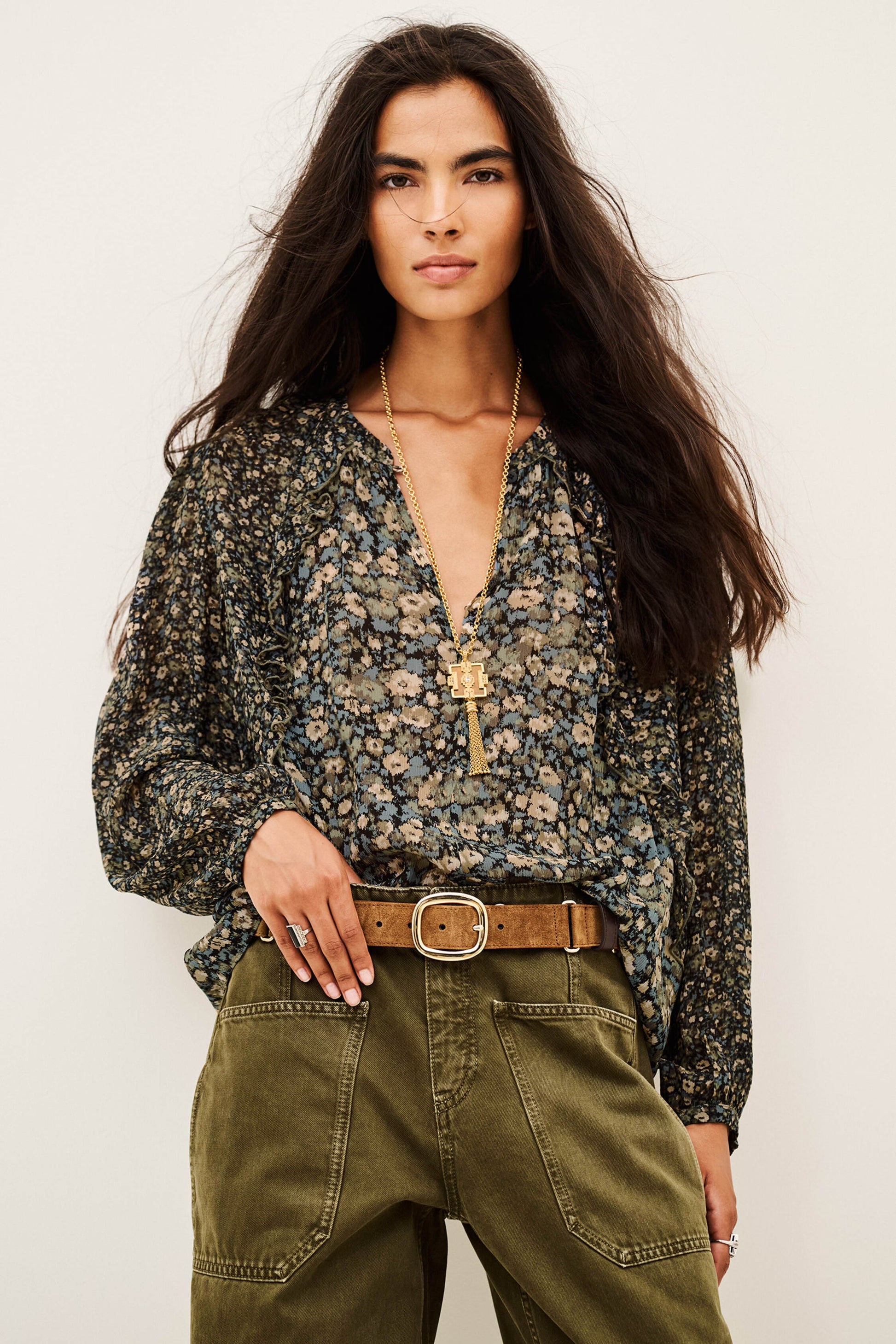 A woman modeling a long-sleeve floral blouse with a deep V-neck and a gold cross pendant, paired with olive green cargo pants and a brown belt, against a plain off-white background.