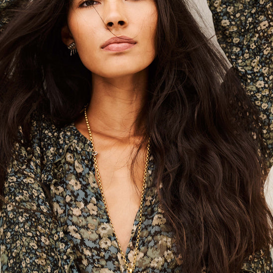 A close-up lifestyle shot of a woman with long, wavy dark hair wearing a dark green and beige floral print top and a gold pendant necklace with a tassel.