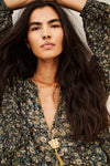 A close-up lifestyle shot of a woman with long, wavy dark hair wearing a dark green and beige floral print top and a gold pendant necklace with a tassel.