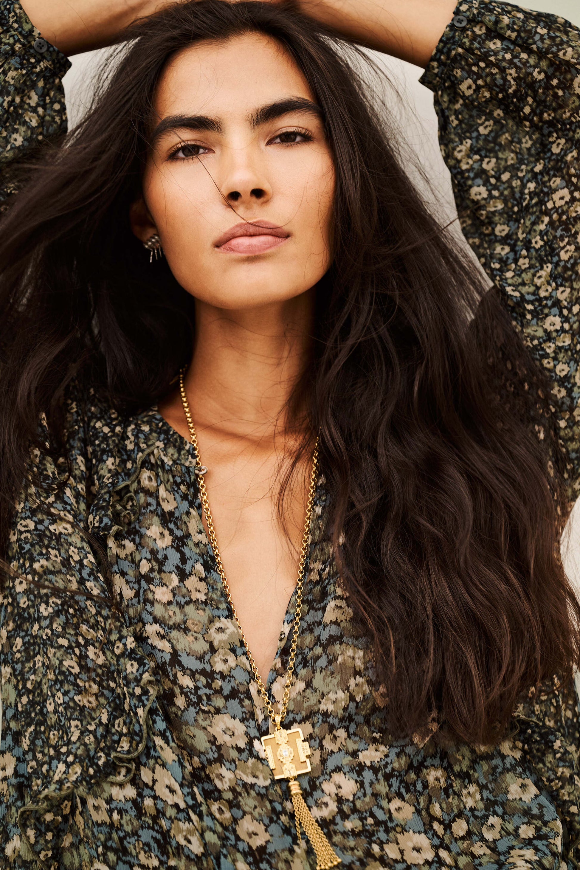 A close-up lifestyle shot of a woman with long, wavy dark hair wearing a dark green and beige floral print top and a gold pendant necklace with a tassel.