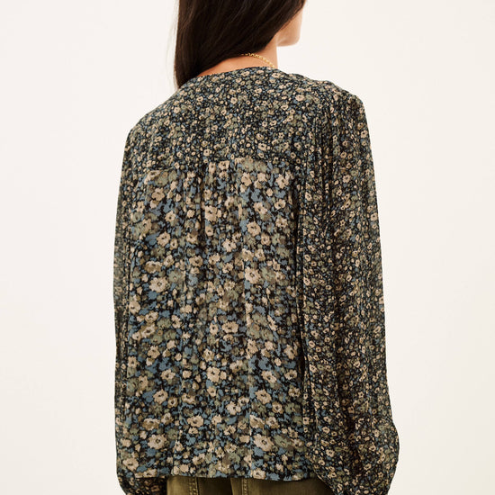 A woman shown from the back wearing a long-sleeve blouse with a dense small floral print in beige, blue, and green tones, paired with olive green pants, against a plain off-white background.