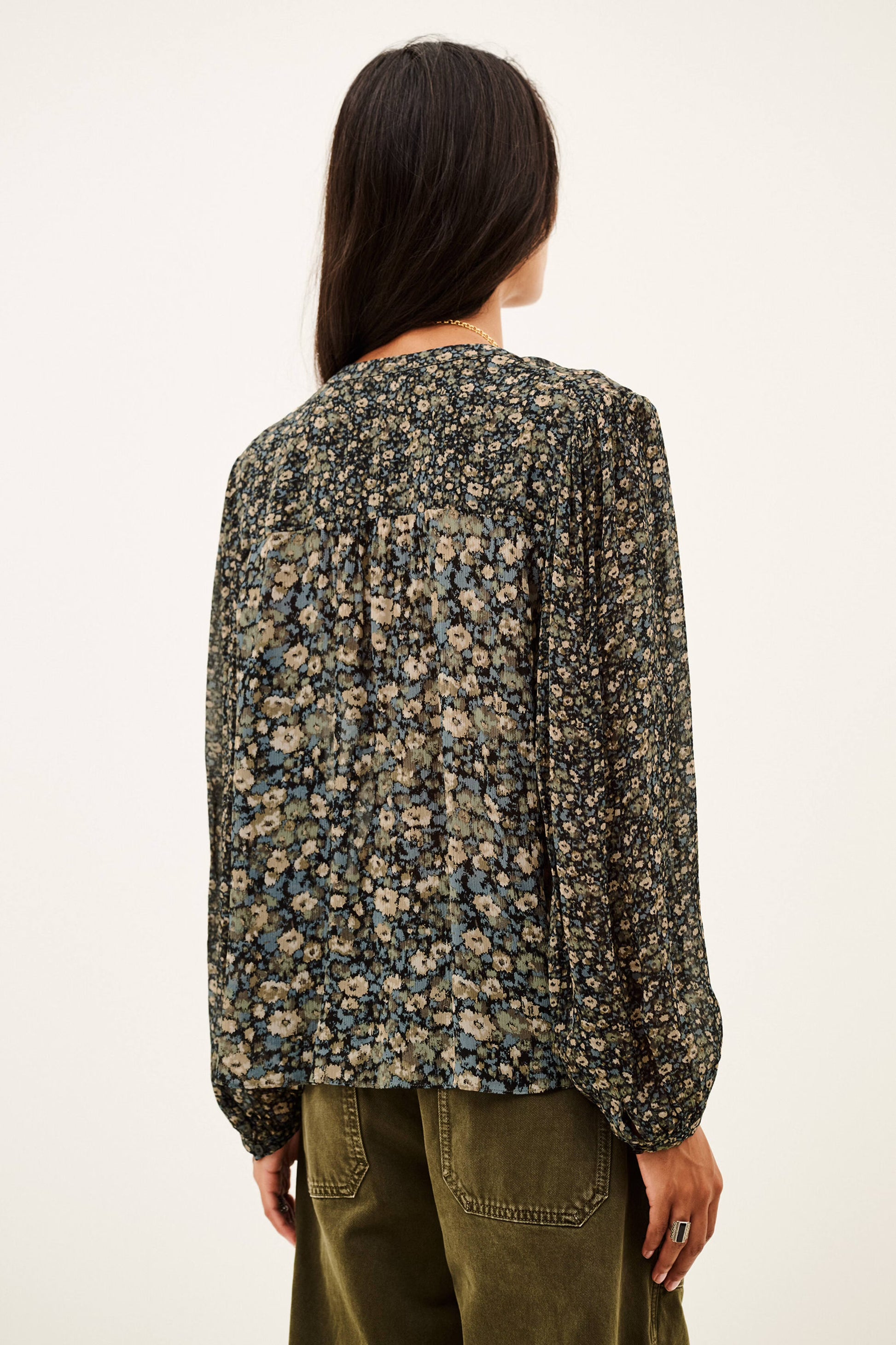 A woman shown from the back wearing a long-sleeve blouse with a dense small floral print in beige, blue, and green tones, paired with olive green pants, against a plain off-white background.