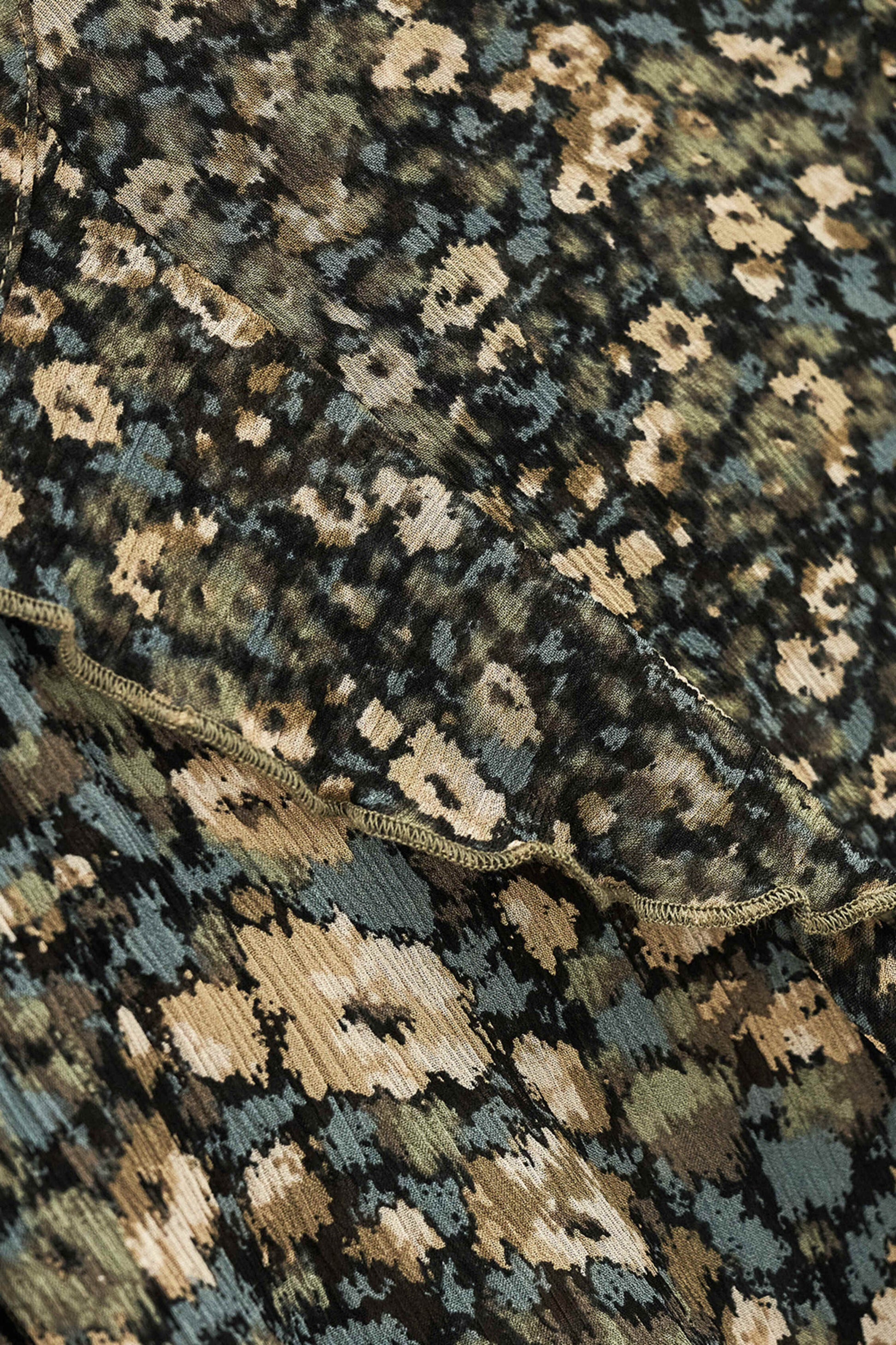 Close-up of a patterned textile with an abstract floral or ikat-style print in black, beige, muted blue-gray, and olive tones, with a scalloped edge visible across the middle.