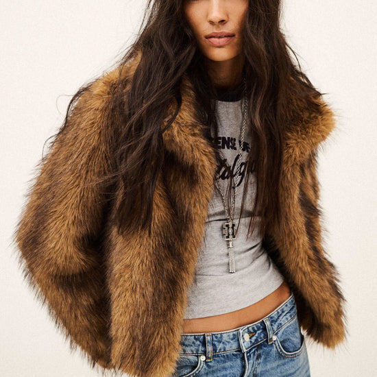 A woman modeling a brown faux fur cropped jacket over a gray graphic crop top and blue jeans, photographed against a plain off-white studio background.
