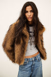 A woman modeling a brown faux fur cropped jacket over a gray graphic crop top and blue jeans, photographed against a plain off-white studio background.