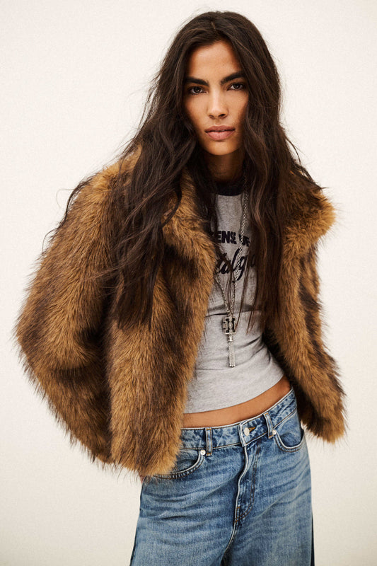 A woman modeling a brown faux fur cropped jacket over a gray graphic crop top and blue jeans, photographed against a plain off-white studio background.