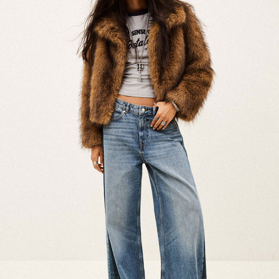 A model wearing a brown faux fur cropped jacket, a light gray graphic crop top, and wide-leg blue denim jeans, standing against a plain off-white studio background.