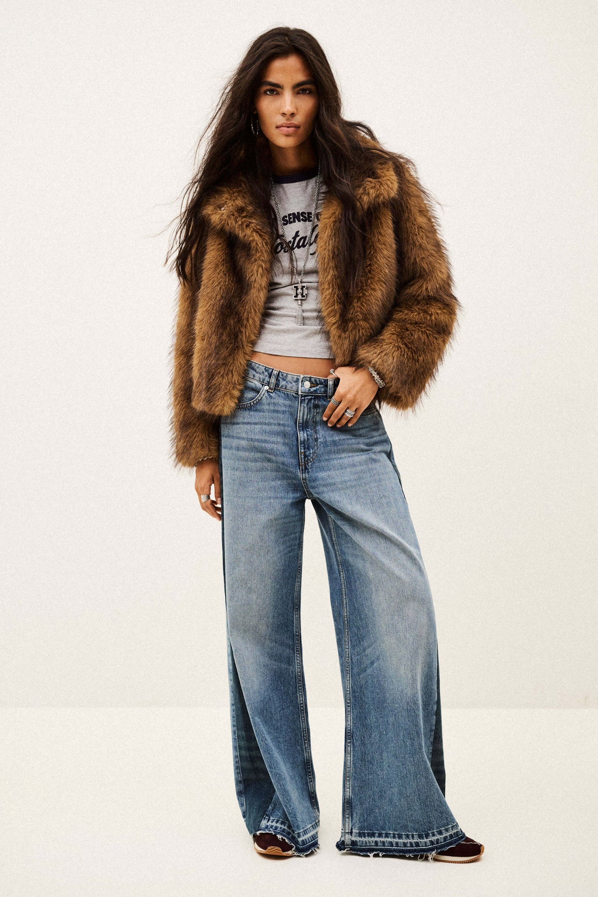 A model wearing a brown faux fur cropped jacket, a light gray graphic crop top, and wide-leg blue denim jeans, standing against a plain off-white studio background.