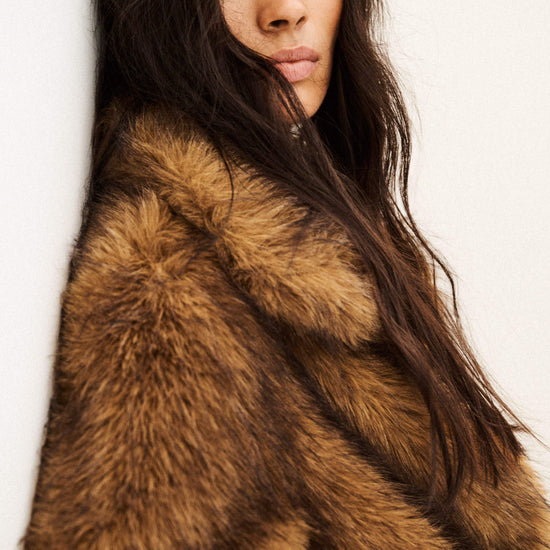 A close-up portrait of a woman wearing a luxurious brown and tan fur coat against a plain off-white background.
