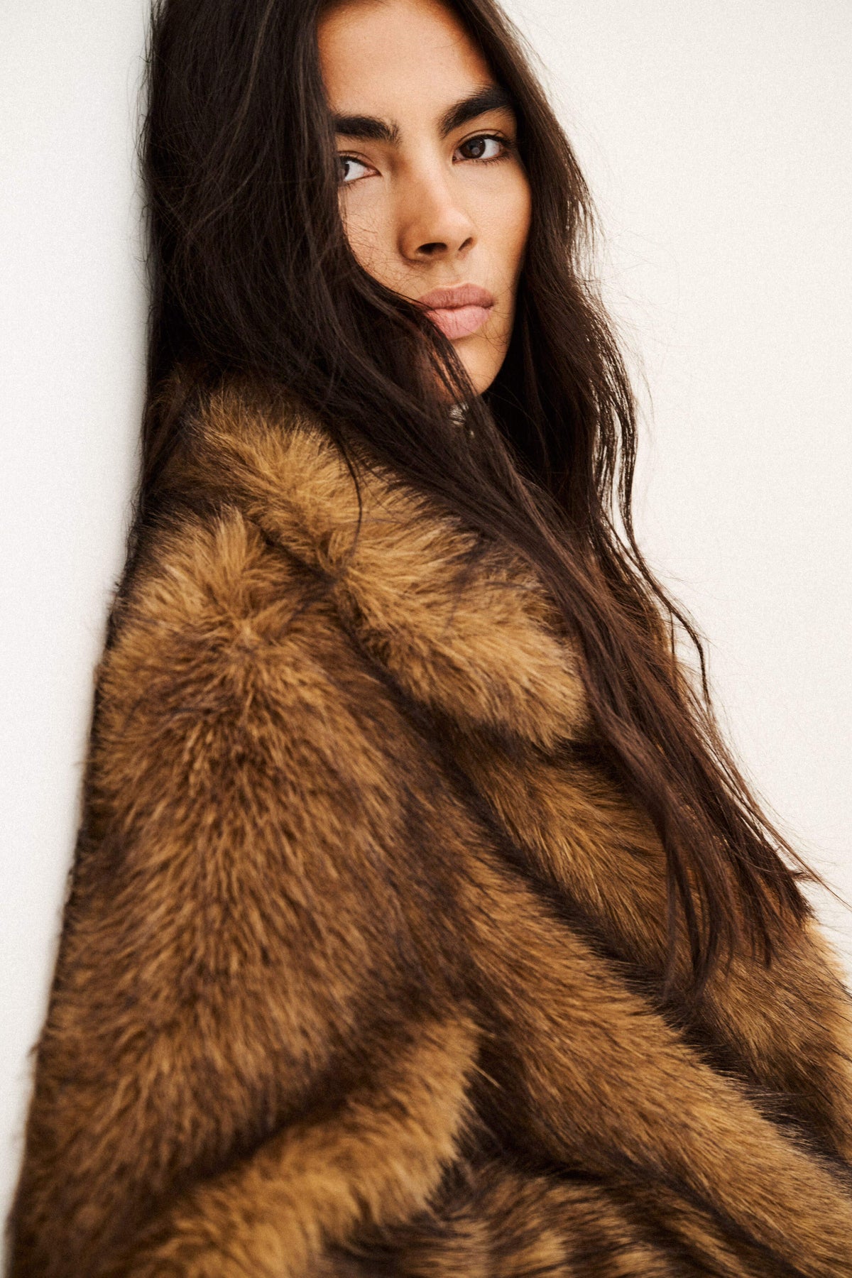 A close-up portrait of a woman wearing a luxurious brown and tan fur coat against a plain off-white background.