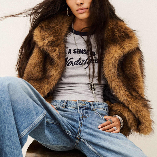 A woman seated on a stool wearing a brown faux-fur jacket over a gray graphic T-shirt and light blue wide-leg jeans, photographed against a plain light background.