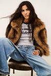 A woman seated on a stool wearing a brown faux-fur jacket over a gray graphic T-shirt and light blue wide-leg jeans, photographed against a plain light background.