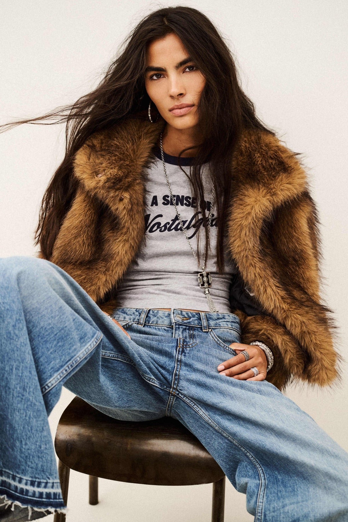 A woman seated on a stool wearing a brown faux-fur jacket over a gray graphic T-shirt and light blue wide-leg jeans, photographed against a plain light background.