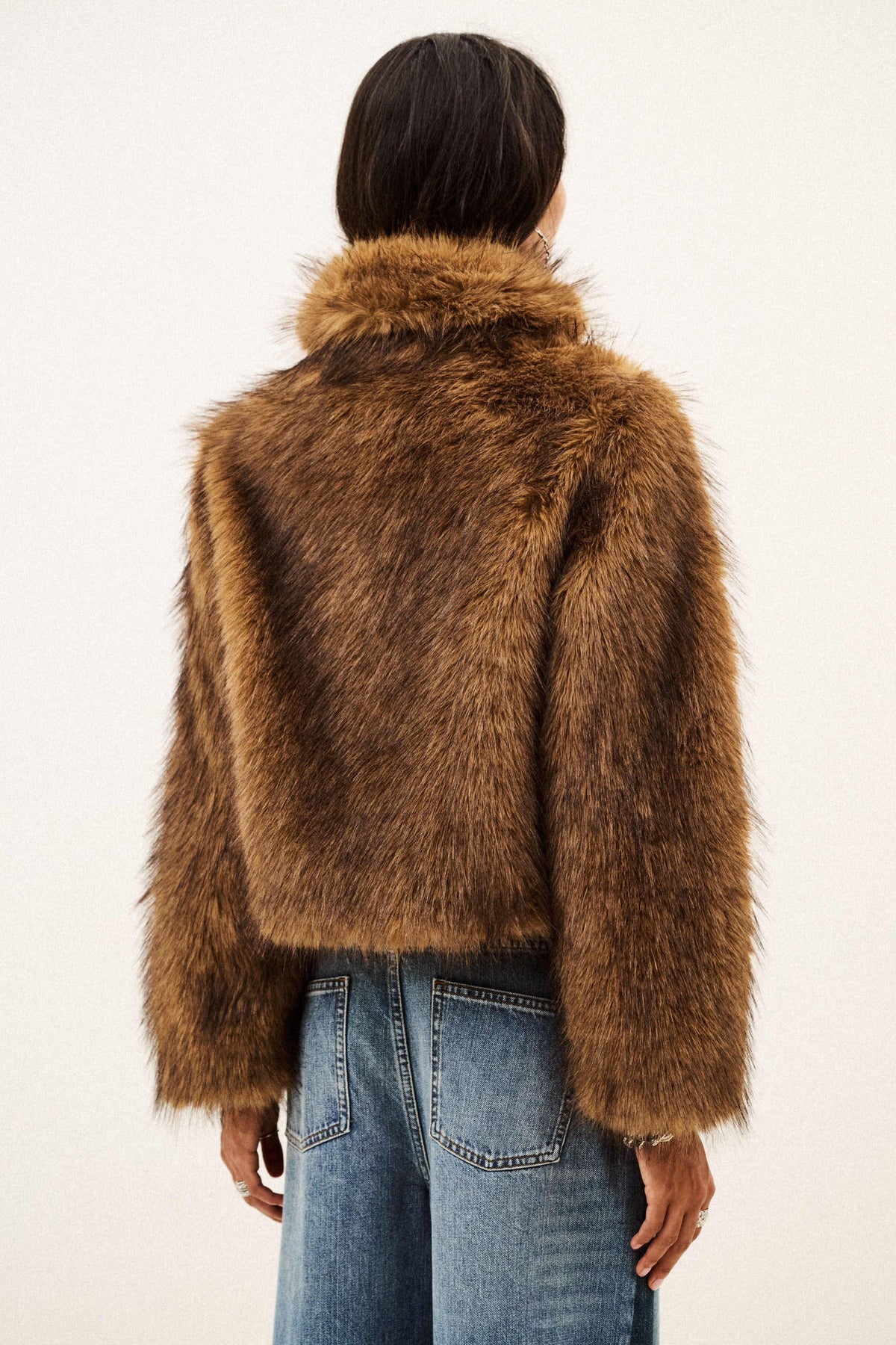 A model viewed from behind wearing a cropped brown faux fur jacket with a wide collar and blue jeans, set against a plain off-white background.