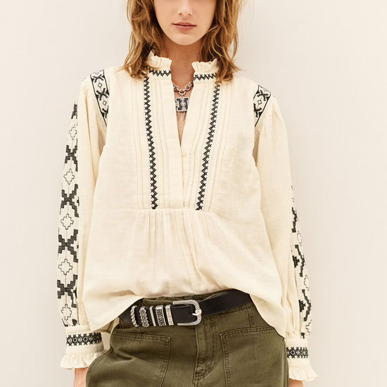 A woman modeling a cream-colored long-sleeve blouse with black geometric embroidery on the sleeves and vertical trim on the front, paired with olive green pants and a black belt.