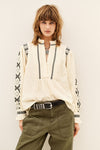 A woman modeling a cream-colored long-sleeve blouse with black geometric embroidery on the sleeves and vertical trim on the front, paired with olive green pants and a black belt.