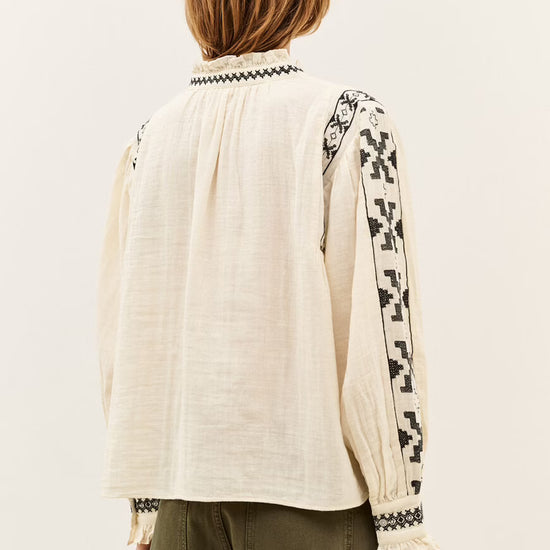 A woman viewed from the back wearing a cream-colored linen blouse with black geometric embroidery on the sleeves and collar, paired with olive green pants, against a plain off-white background.