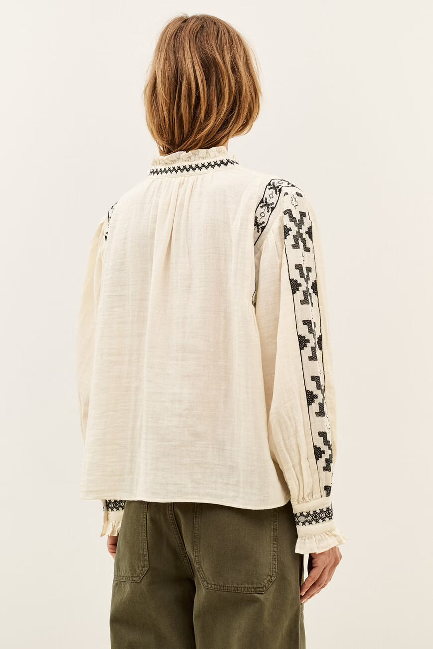 A woman viewed from the back wearing a cream-colored linen blouse with black geometric embroidery on the sleeves and collar, paired with olive green pants, against a plain off-white background.
