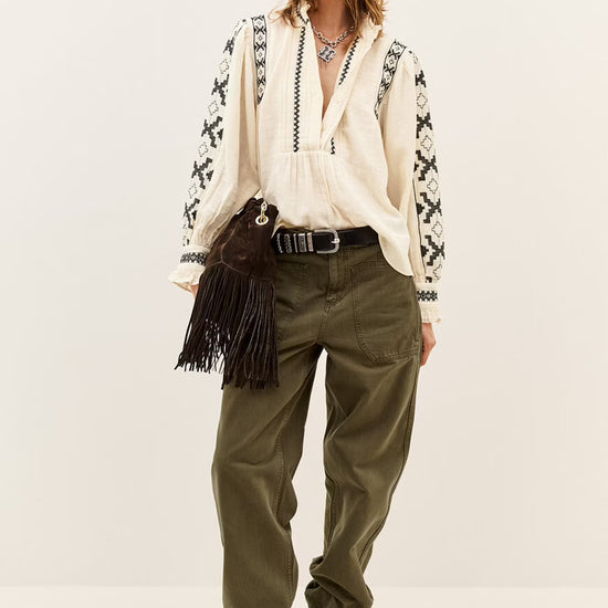 A model stands against a plain light beige studio background wearing a bohemian-style outfit consisting of an off-white embroidered cardigan, a cream blouse, olive green pants, brown suede boots, and a dark brown fringe bag.