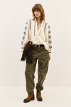 A model stands against a plain light beige studio background wearing a bohemian-style outfit consisting of an off-white embroidered cardigan, a cream blouse, olive green pants, brown suede boots, and a dark brown fringe bag.