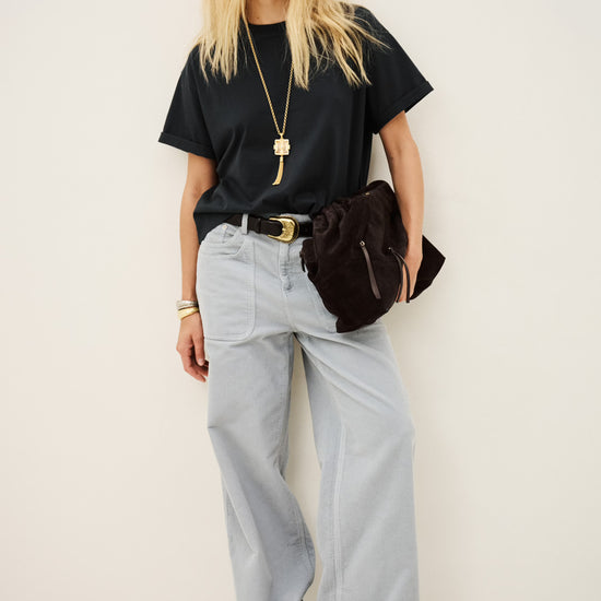 A blonde model stands against a plain off-white studio wall wearing an oversized black t-shirt, light gray wide-leg jeans, a black belt with a gold buckle, and carrying a dark brown shoulder bag.