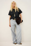 A blonde model stands against a plain off-white studio wall wearing an oversized black t-shirt, light gray wide-leg jeans, a black belt with a gold buckle, and carrying a dark brown shoulder bag.