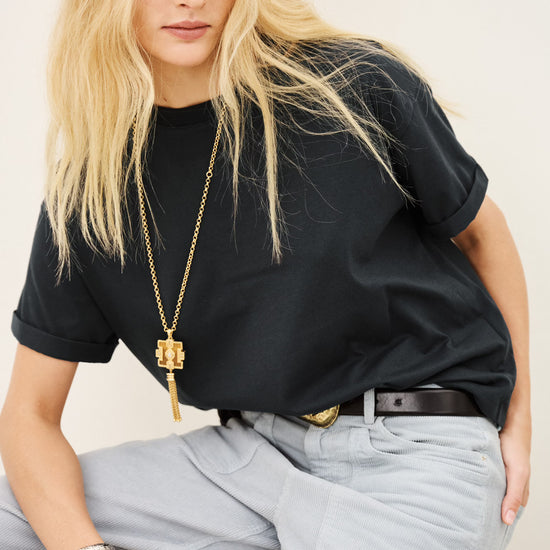 A blonde woman wearing a black short-sleeve t-shirt, light gray wide-leg jeans, a black belt, and gold jewelry, sitting on a wooden block against a plain off-white background.