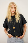A blonde woman wearing a black short-sleeve t-shirt, light gray jeans, a gold pendant necklace, and gold bracelets, posing against a light off-white background.
