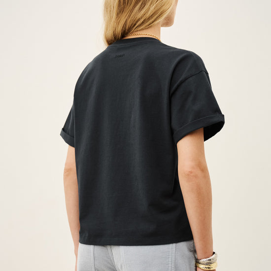 A person shown from the back wearing a black short-sleeve crewneck t-shirt and light gray pants, photographed against a plain off-white studio background.