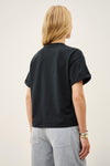A person shown from the back wearing a black short-sleeve crewneck t-shirt and light gray pants, photographed against a plain off-white studio background.