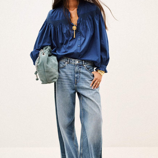A model wearing a royal blue satin blouse with voluminous sleeves, wide-leg light-wash blue jeans, and carrying a small sage green drawstring bag against a plain off-white studio background.