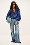 A model wearing a royal blue satin blouse with voluminous sleeves, wide-leg light-wash blue jeans, and carrying a small sage green drawstring bag against a plain off-white studio background.