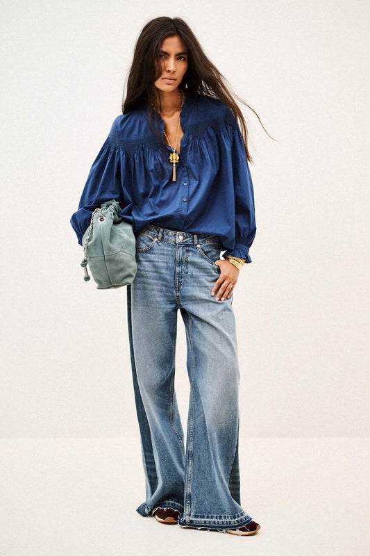 A model wearing a royal blue satin blouse with voluminous sleeves, wide-leg light-wash blue jeans, and carrying a small sage green drawstring bag against a plain off-white studio background.