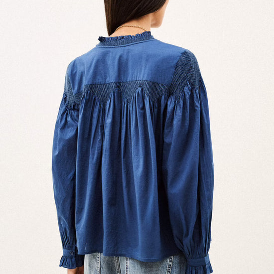 A woman viewed from the back wearing a blue long-sleeve blouse with gathered detailing and ruffled trim, paired with light blue jeans, against a plain off-white background.
