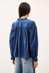 A woman viewed from the back wearing a blue long-sleeve blouse with gathered detailing and ruffled trim, paired with light blue jeans, against a plain off-white background.