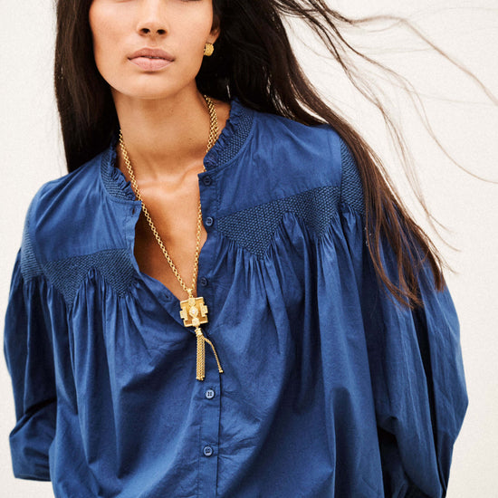 A woman modeling a blue long-sleeve blouse with gathered puff sleeves, a split neckline, and subtle darker blue embroidery details, paired with blue jeans and a gold pendant necklace against a plain light background.