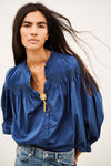 A woman modeling a blue long-sleeve blouse with gathered puff sleeves, a split neckline, and subtle darker blue embroidery details, paired with blue jeans and a gold pendant necklace against a plain light background.