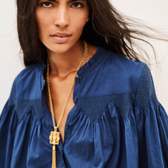 A woman wearing a blue blouse and a gold pendant necklace, photographed against a plain light background.